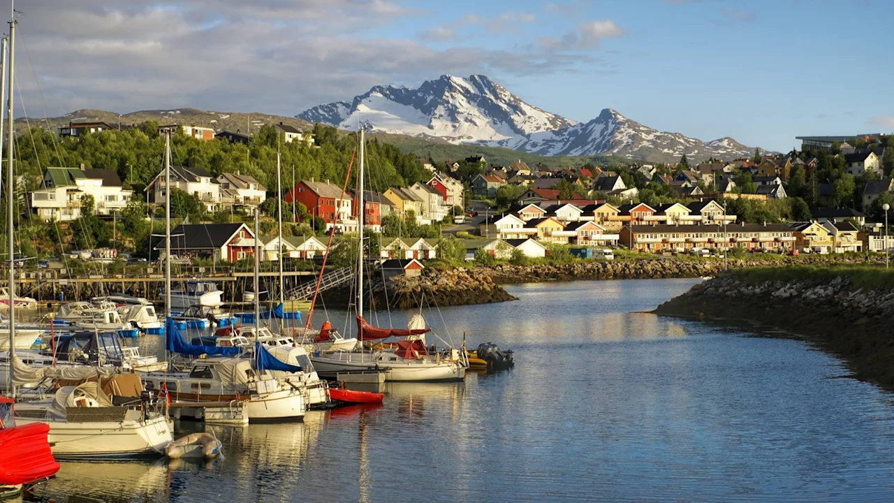 Sailboats rest in a marina, surrounded by colorful houses and lush greenery with snow-capped mountains in the background under a blue sky.