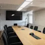 Et langt trækonferencebord med sorte stole står foran en vægmonteret skærm. Rummet er godt oplyst med loftlamper, trægulv og minimal udsmykning.