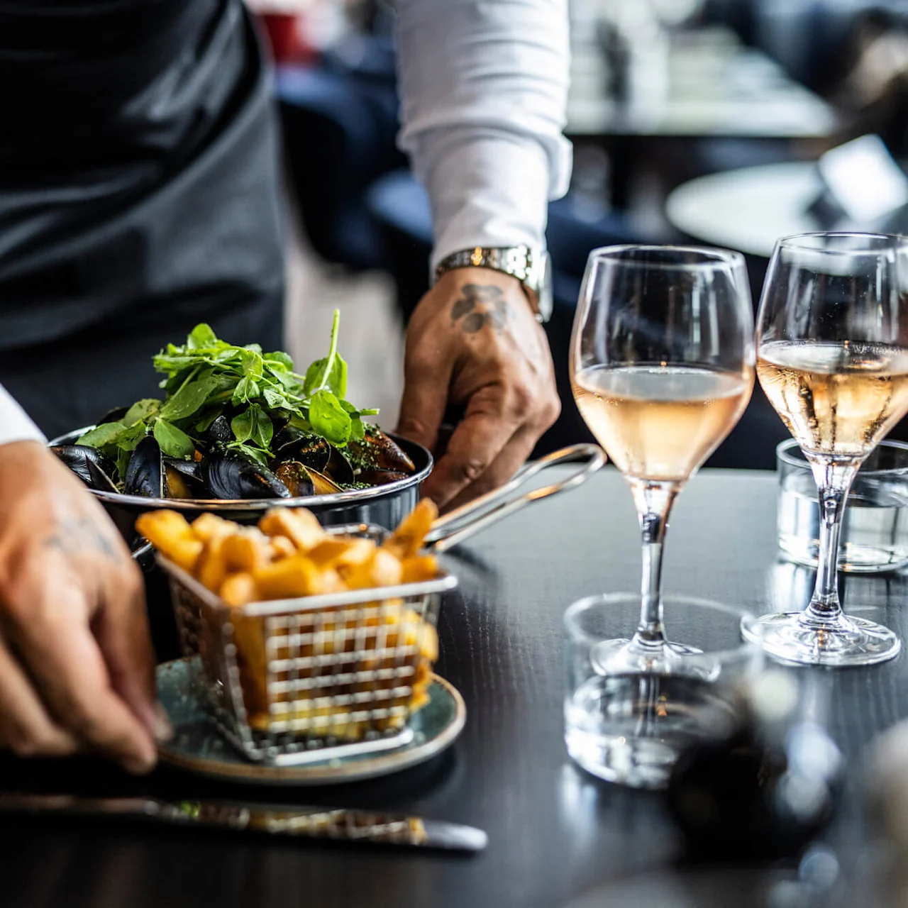 En servitör serverar musslor med örter och pommes frites vid ett bord, tillsammans med två vinglas, i en restaurangmiljö.
