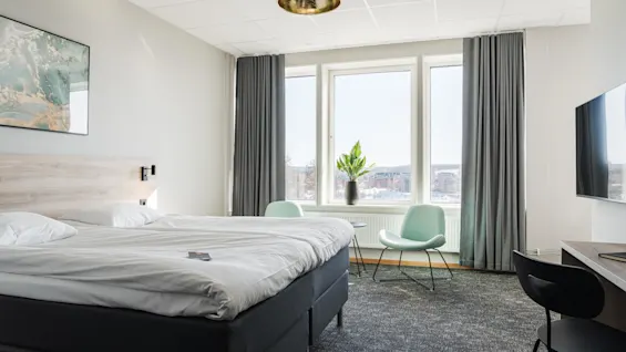 En prydligt bäddad säng står i ett modernt hotellrum. Två stolar och ett bord vetter mot ett stort fönster med utsikt över staden. Mjuk belysning och konstverk förhöjer miljön.