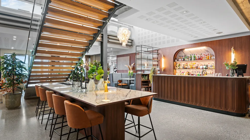 Et marmorbord med brune stoler omgir en trapp, dekorert med planter og vaser. I nærheten er en trebardisk med opplyste hyller som viser ulike flasker.