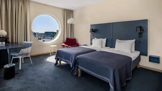 A bright hotel room with twin beds, a comfortable workspace, and a large circular window showcasing a city view.