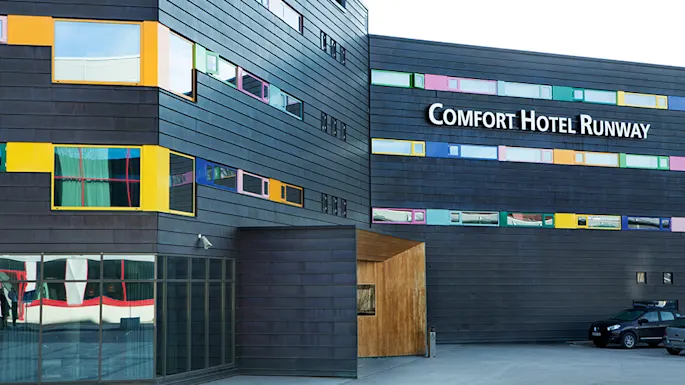 A modern hotel building has colorful windows set in dark, paneled walls. The sign reads "COMFORT HOTEL RUNWAY." A parked car is visible in front.
