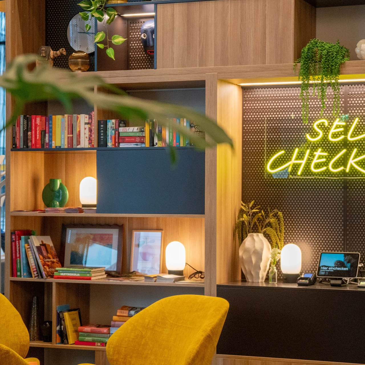 Bookshelf with neon sign “SELF CHECK-IN” on display. Nearby are small devices and decorative plants. Bright seating and books fill warm, inviting space.