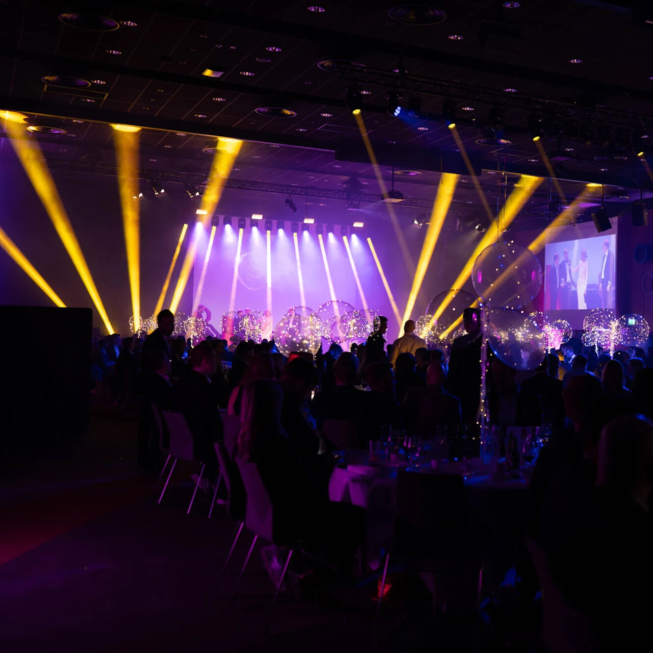 Colorful spotlights illuminate guests sitting at round tables in a grand hall during an event. Large screens display people on stage, enhancing the festive atmosphere.