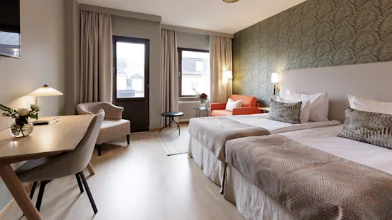 Två enkelsängar med ljusfärgade lakan i ett välbelyst hotellrum med skrivbord, fåtölj och dekorativ tapet.