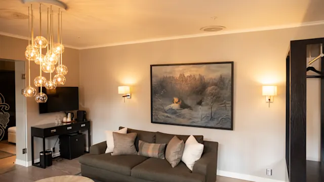 En koselig stue med en grå sofa dekorert med puter, plassert mot en lys vegg med et stort vinterlandskapsmaleri. Myk belysning gis av en moderne lysekrone.