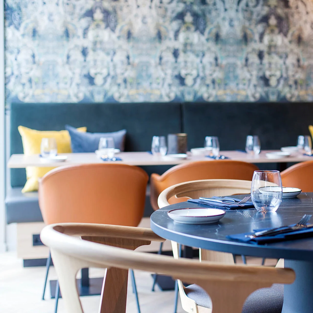 Et rundt spisebord er dekket med glass, tallerkener og bestikk. Det er på en stilfull restaurant dekorert med mønstret tapet og polstrede stoler.