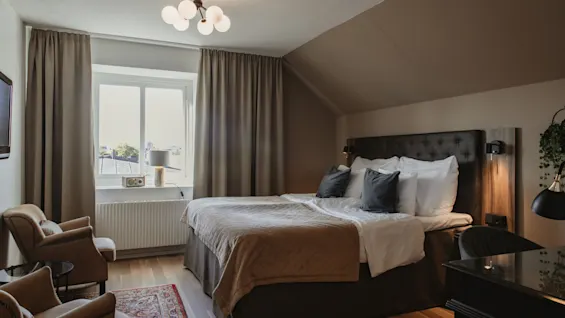 Viihtyisä hotellihuone, jossa on suuri sänky, kaksi nojatuolia ja työpöytä, kylpee luonnonvalossa ikkunasta.