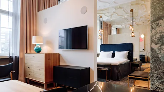 A flat-screen TV is mounted on a wall divider in a modern bedroom. It faces a wooden dresser with a lamp and nearby a bed with pendant lights.