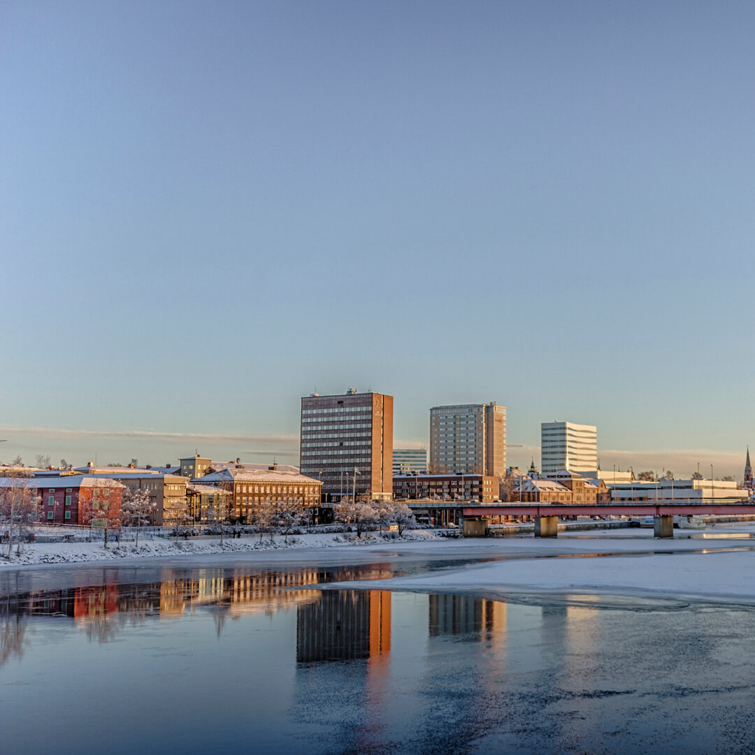 En bysilhuet med bygninger, der reflekteres på delvist frossent flodvand under en klar blå himmel ved daggry eller skumring.