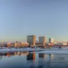 En byskyline med bygninger som reflekteres i delvis frosset elvevann under en klar blå himmel ved daggry eller skumring.