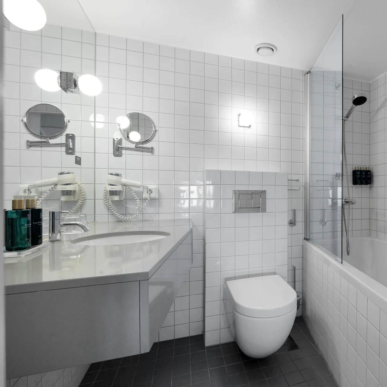 Et hvitt flislagt bad med vegghengt toalett, vask med toalettsaker, badekar med dusjhode og speil med spotlights. En hårføner og et håndkle er synlige.