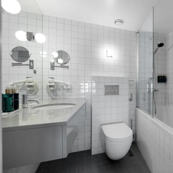 Et hvitt flislagt bad med vegghengt toalett, vask med toalettsaker, badekar med dusjhode og speil med spotlights. En hårføner og et håndkle er synlige.