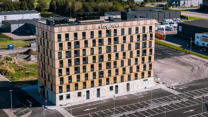Et høyt, rektangulært hotell ved navn "The Urban House" står på en stor parkeringsplass, med et industrielt landskap med spredte lagerbygninger og grønne jorder under en delvis skyet himmel.