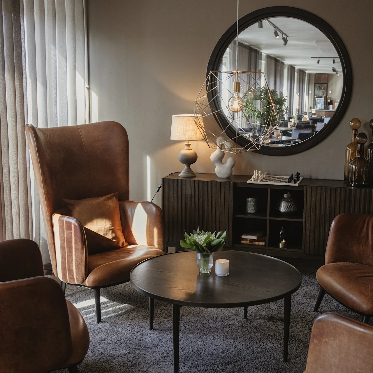 Four brown leather armchairs encircle a round coffee table with a plant and candles, situated in a cozy, warmly lit living room featuring a mirror, lamp, and decorative sculptures.