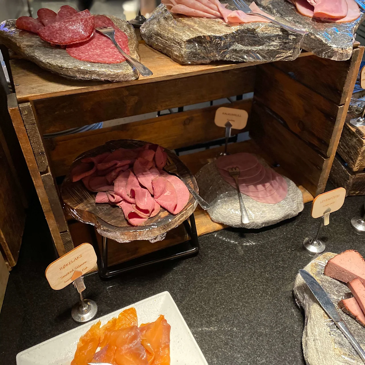 Various sliced meats, including salami, ham, pastrami, and smoked salmon, are displayed on wooden platters and stone slabs in a buffet setting. Text: "Salami," "HOTELLSKINKE," "ROASTBIFF," "KAJARERULL," "RØKELAKS," "LØREROFPSTI."