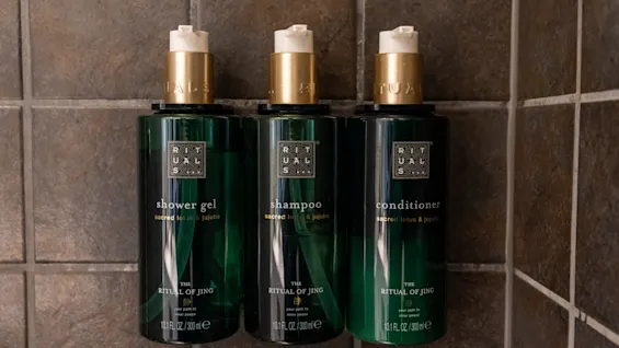 Tre grønne flasker mærket "shower gel", "shampoo" og "conditioner" monteret på en flisevæg. Tekst: "sacred lotus & jujube", "THE RITUAL OF JING", "10.1 FL. OZ./300 ml".
