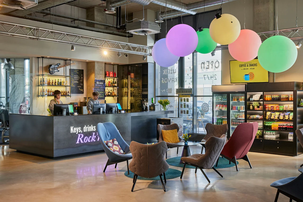 Reception desk with two attendants assisting guests, surrounded by colorful chairs and balloons. Nearby, shelves display snacks and drinks. Visible text: “Keys, drinks Rock’n”, “COFFEE & SNACKS 09.00 - 19.00”.