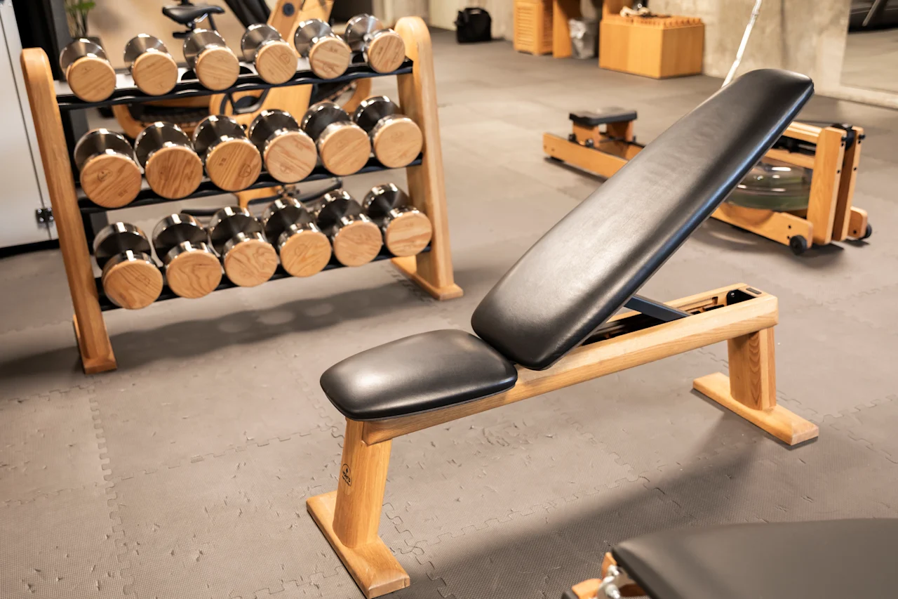 Träningsbänk på ett golvgymgolv; i närheten håller ett träställ olika hantlar, vilket tyder på en fitnessstudio-miljö med ytterligare utrustning i bakgrunden.