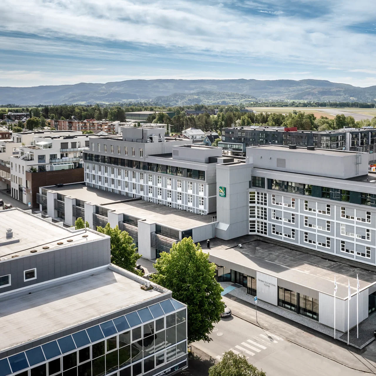 En stor, moderne bygning med hvid og grå facade ligger blandt en urban omgivelser med andre bygninger og fjerne bjerge. Tre hvide flag er ved indgangen.