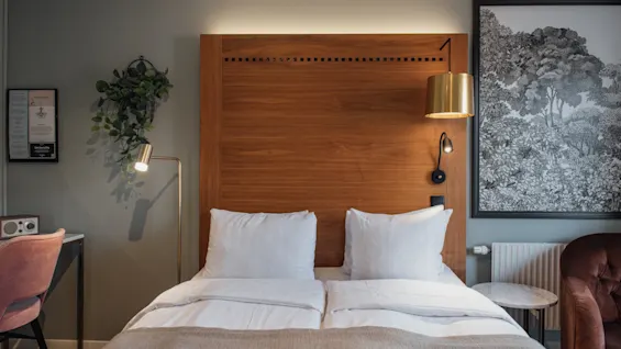 Et hotelværelse med en seng, sengegavl i træ og blød belysning. En plante, skrivebord og indrammet kunst skaber en hyggelig atmosfære.