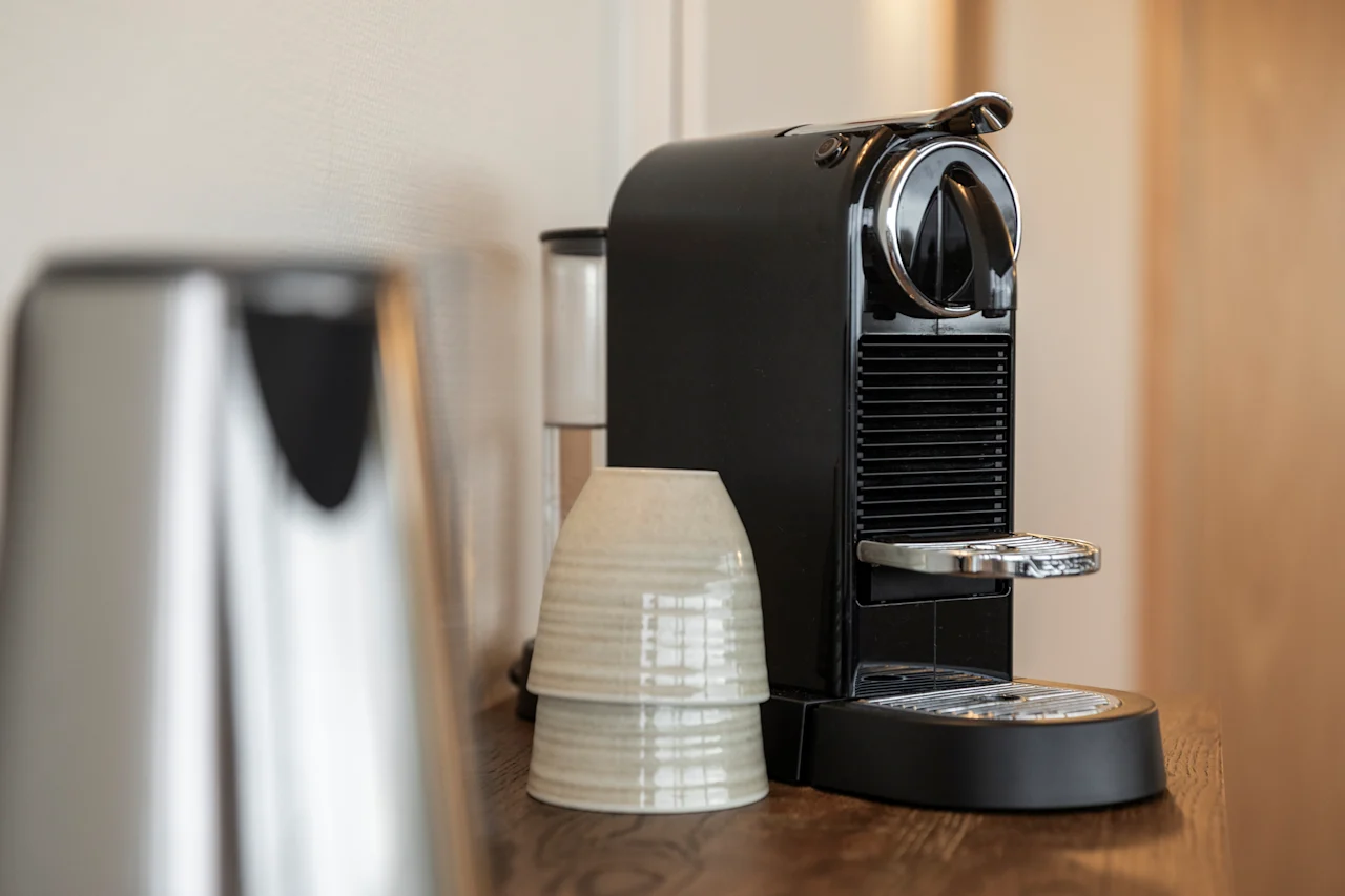 A black coffee machine, stacked cups, and a kettle on a wooden surface, suggesting a hot drink is about to be made.