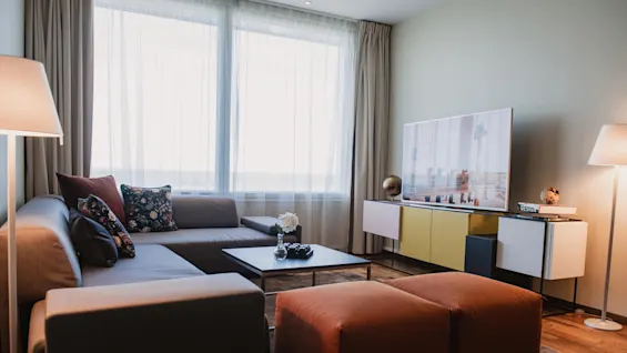 A cozy living room features a grey sectional sofa with decorative pillows, facing a TV on a modern console. Soft lighting and large windows create a warm atmosphere.