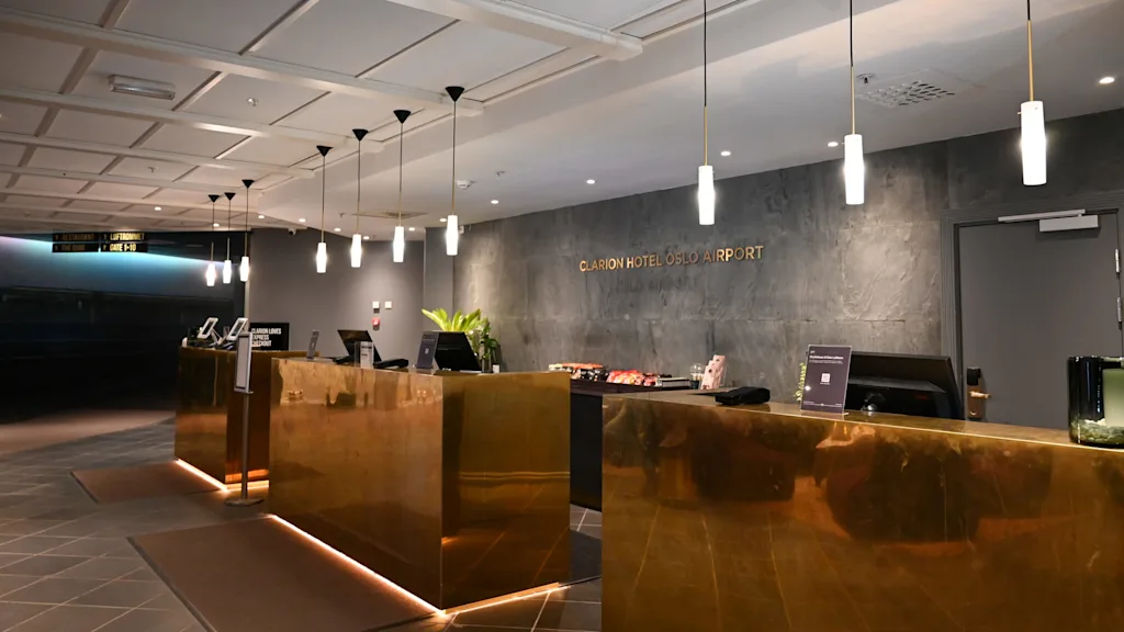 Reception med messingdiske under hængelamper, et skilt med "Clarion Hotel Oslo Airport" på en struktureret væg og moderne indretning inklusive planter. Skilte viser retning til faciliteter.