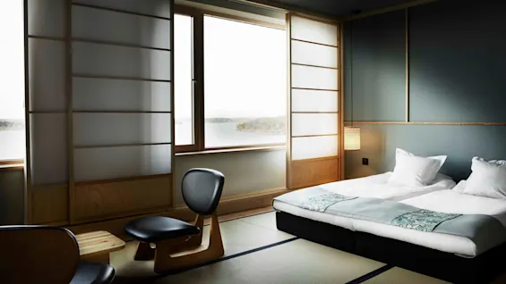 Et minimalistisk soverom med en lav plattformseng, et moderne stolsett, fredelig innsjøutsikt og mykt, diffust lys som filtreres gjennom et stort papirpanelvindu.