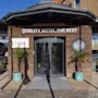 En ingång av glas märkt "QUALITY HOTEL THE REEF" med sido-skyltar "SociAL" och "Reef"; omgiven av växter i krukor på en skuggig uteplats med sittplatser utomhus.