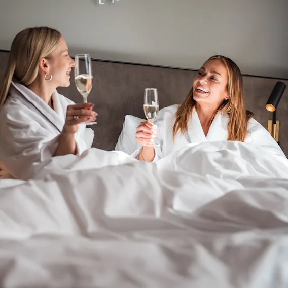 Kaksi kylpytakkiin pukeutunutta henkilöä sängyssä, hymyilevät ja kilistävät samppanjalaseilla. Nauttivat rentouttavasta hetkestä.