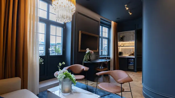Elegant hotel room featuring dark blue walls, stylish leather armchairs, a crystal chandelier, and large windows.