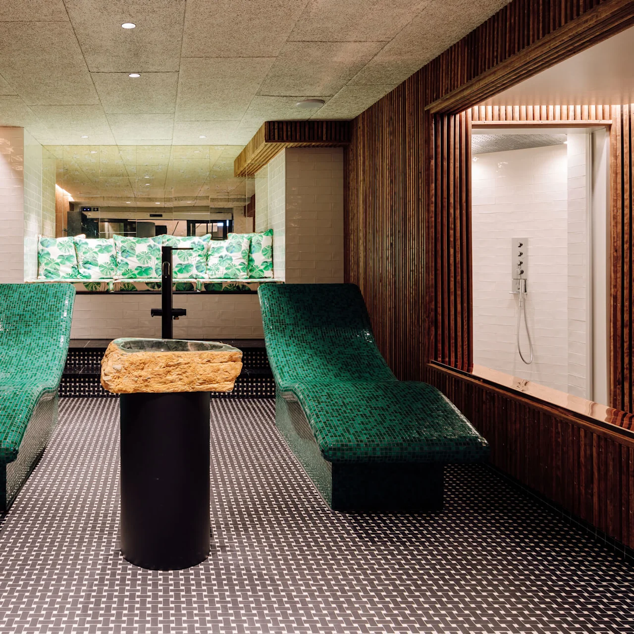Two green mosaic-tiled lounge chairs face each other across a stone-topped pedestal sink in a wood-paneled room with patterned floor tiles. A shower area is visible in the background.