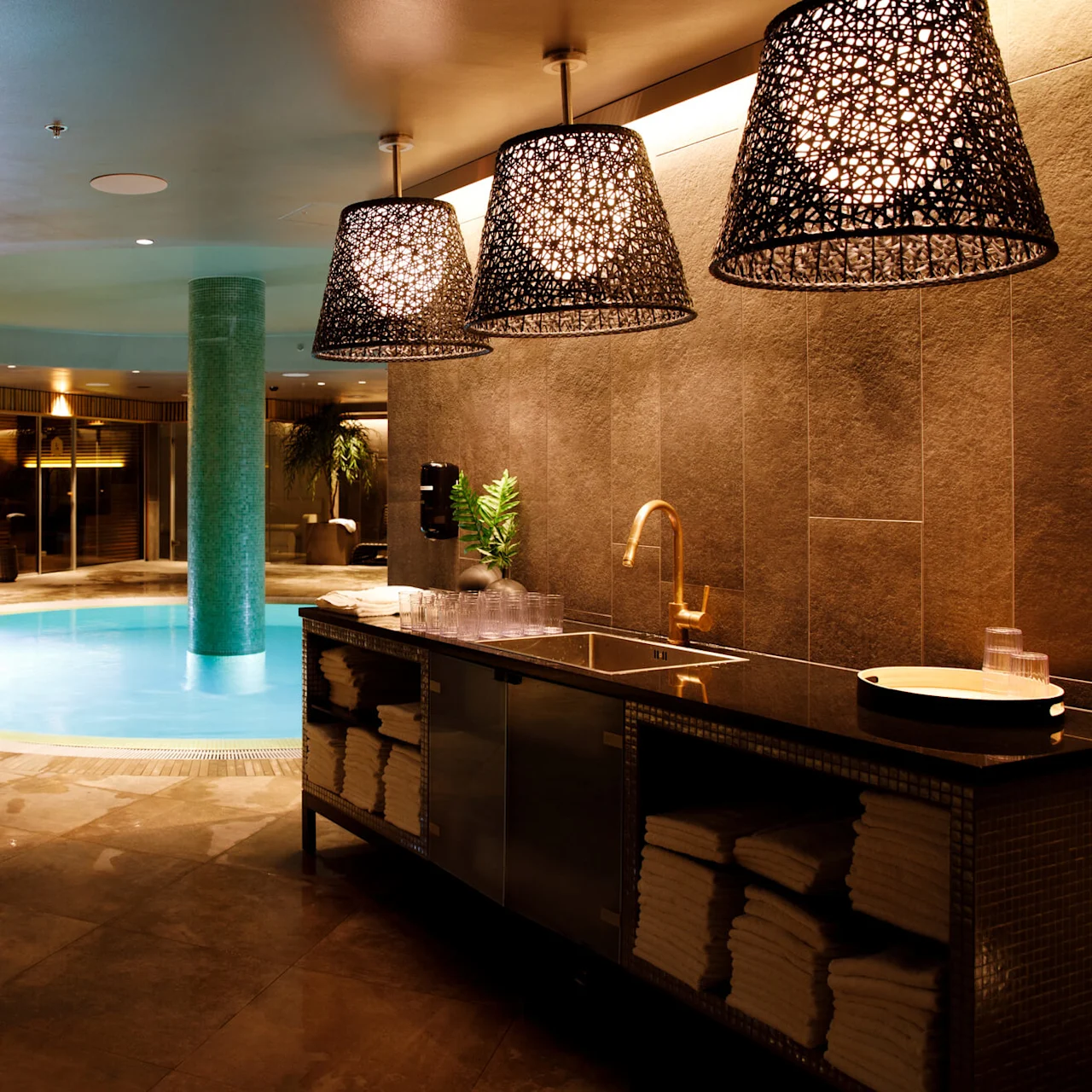Pool with glowing blue water sits under dim, decorative lighting. Nearby, a countertop holds neatly stacked towels and cups, creating a spa-like atmosphere with wooden accents and soft illumination.