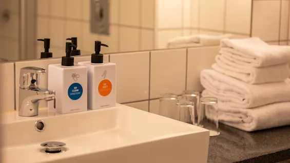 Hvit vask med en kromkran har dispensere merket "GO CLEAN HAND SOAP" og "GO SOFT BODY LOTION." Brettede håndklær og glass på benkeplaten på et flislagt bad.