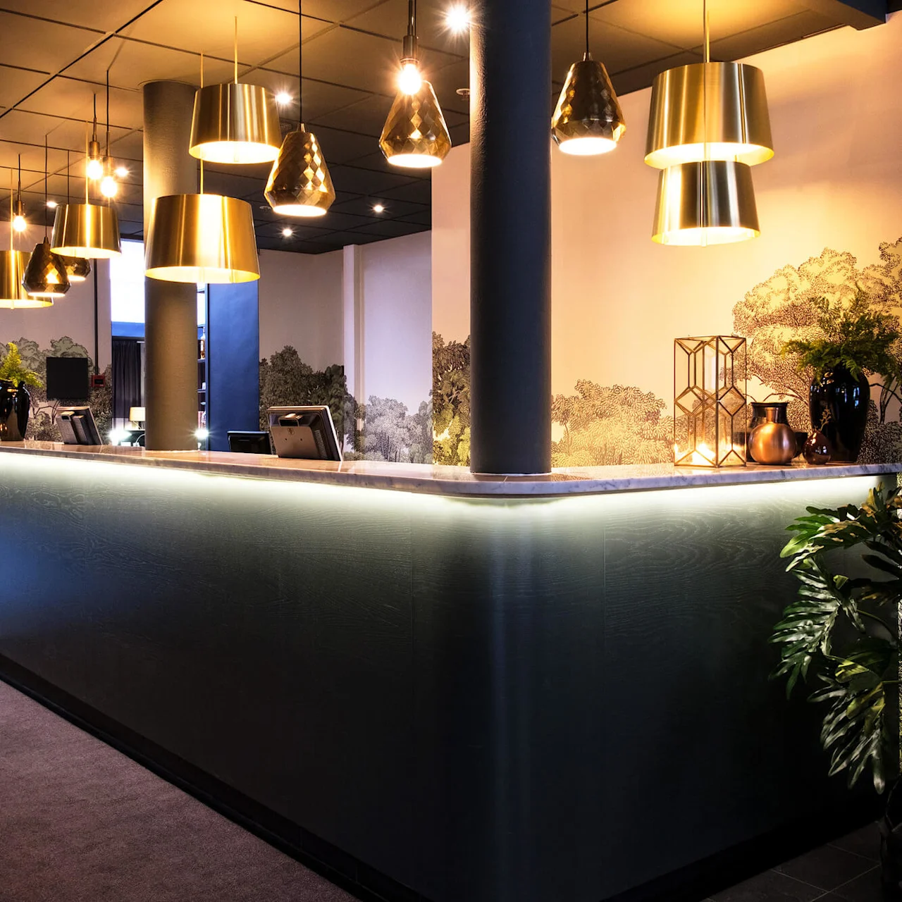 A sleek reception desk with overhead pendant lights in a stylish lobby, featuring decorative plants and a mural.