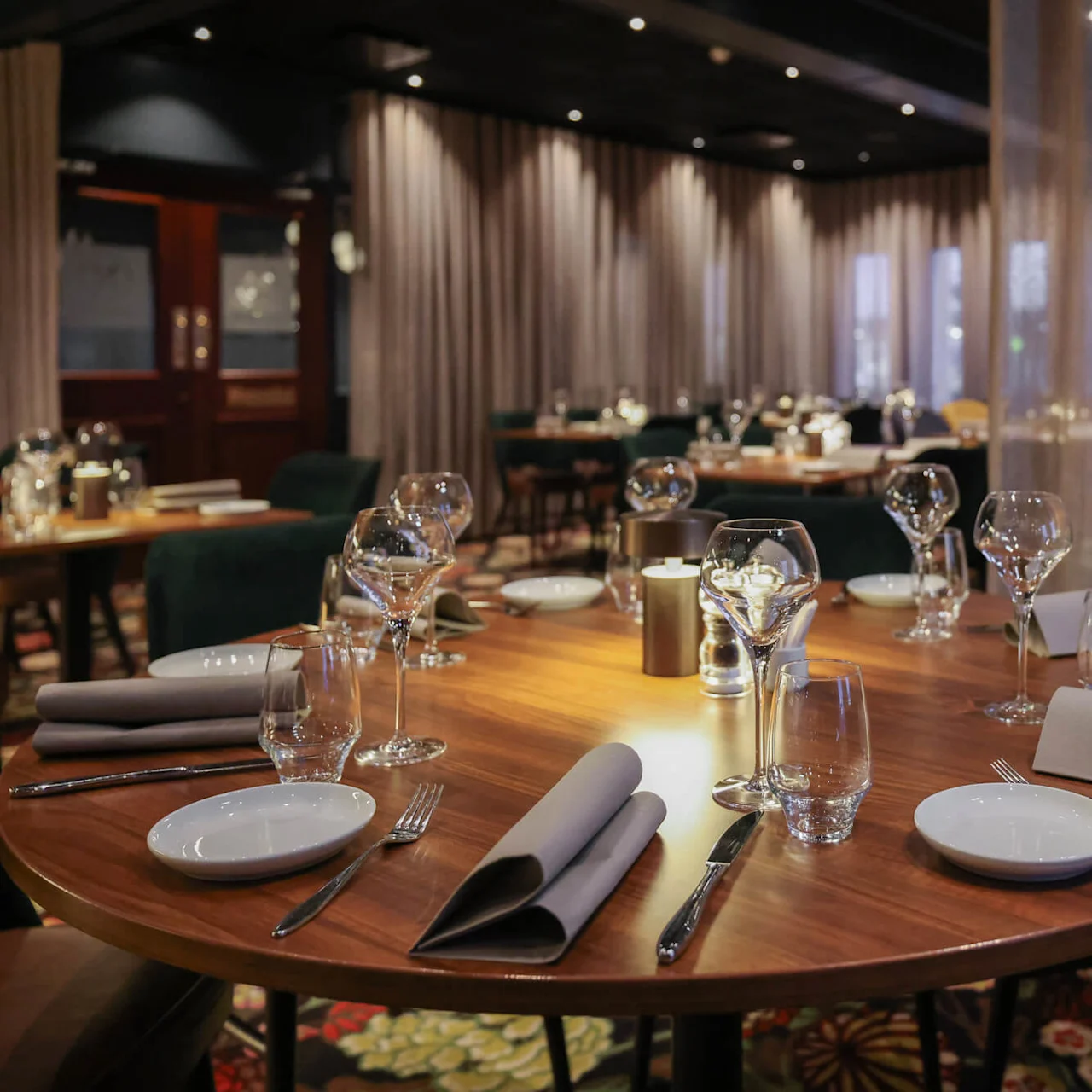 Round wooden table set with wine glasses, plates, and napkins in an elegant, dimly-lit restaurant with upholstered chairs and curtained walls.
