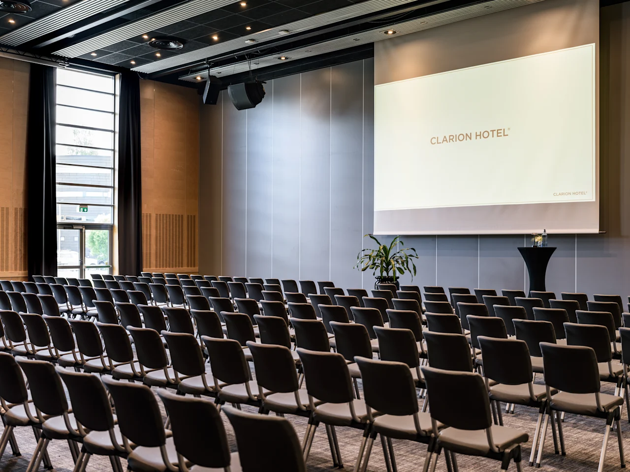 Ett rymligt konferensrum med rader av stolar vända mot en stor skärm som visar CLARION HOTEL. Naturligt ljus fyller rummet.