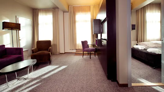 Et moderne hotellrom med lilla og brune møbler, naturlig lys som flommer gjennom gjennomsiktige gardiner, og et synlig soverom.