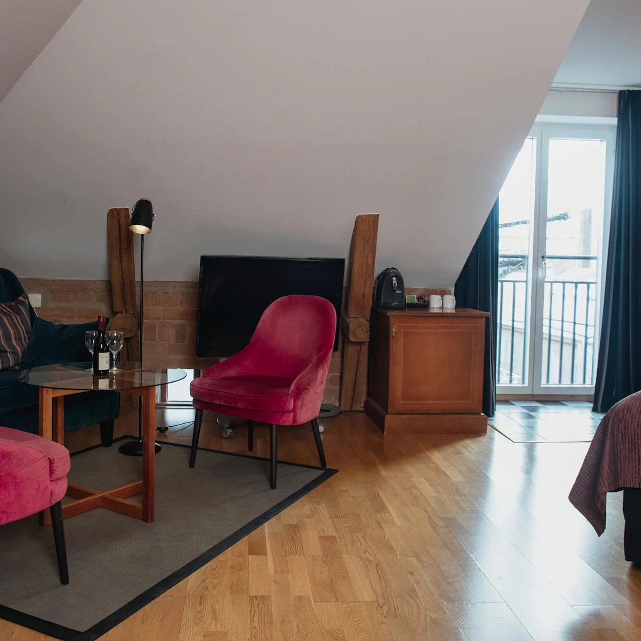 A cozy attic room features a seating area with a blue sofa and pink chairs around a coffee table. A balcony with glass doors provides natural light.