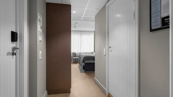A hotel room hallway with a bed and chairs visible through an open doorway. A key card reader is on the left door. Get b