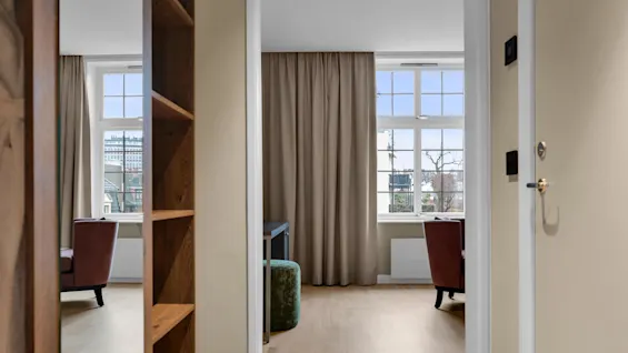 En dörröppning leder till ett ljust hotellrum med stora fönster och gardiner; trägolv sträcker sig genom utrymmet.