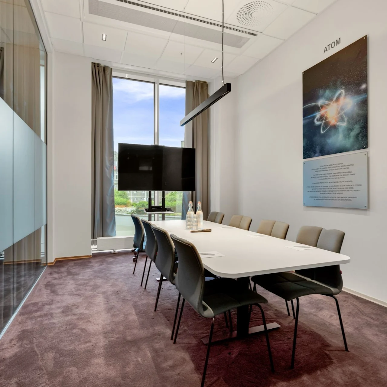 A conference room with a long white table and eight chairs is set for a meeting. A screen and artwork titled "ATOM" are mounted on the wall. Large windows provide natural light.