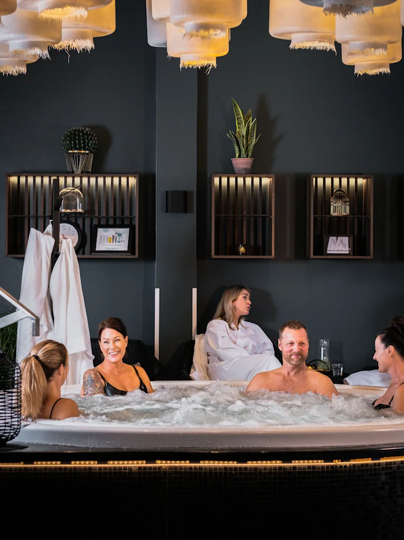 People relaxing and enjoying a bubbly hot tub in a dimly lit spa area with decorative lighting and plants.