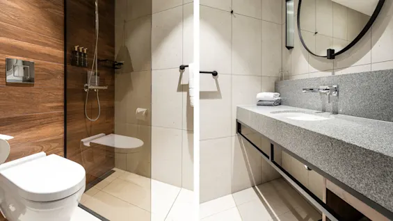 Brusebad og håndvask med moderne design; brusebad med glasvæg og trævæg, håndvask med stort rundt spejl og håndklæder; badeværelse i neutrale toner og minimalistisk indretning.