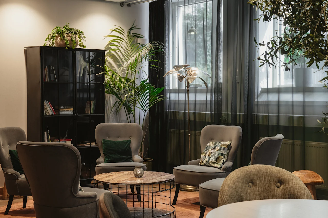 A cozy lounge area at Home Hotel Uppsala with comfortable armchairs, a coffee table, a bookshelf, and lush green plants.