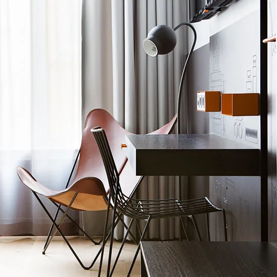 Seng med hvitt sengetøy står ved siden av en moderne stol og skrivebord med lampe i et rom med gjennomsiktige gardiner.