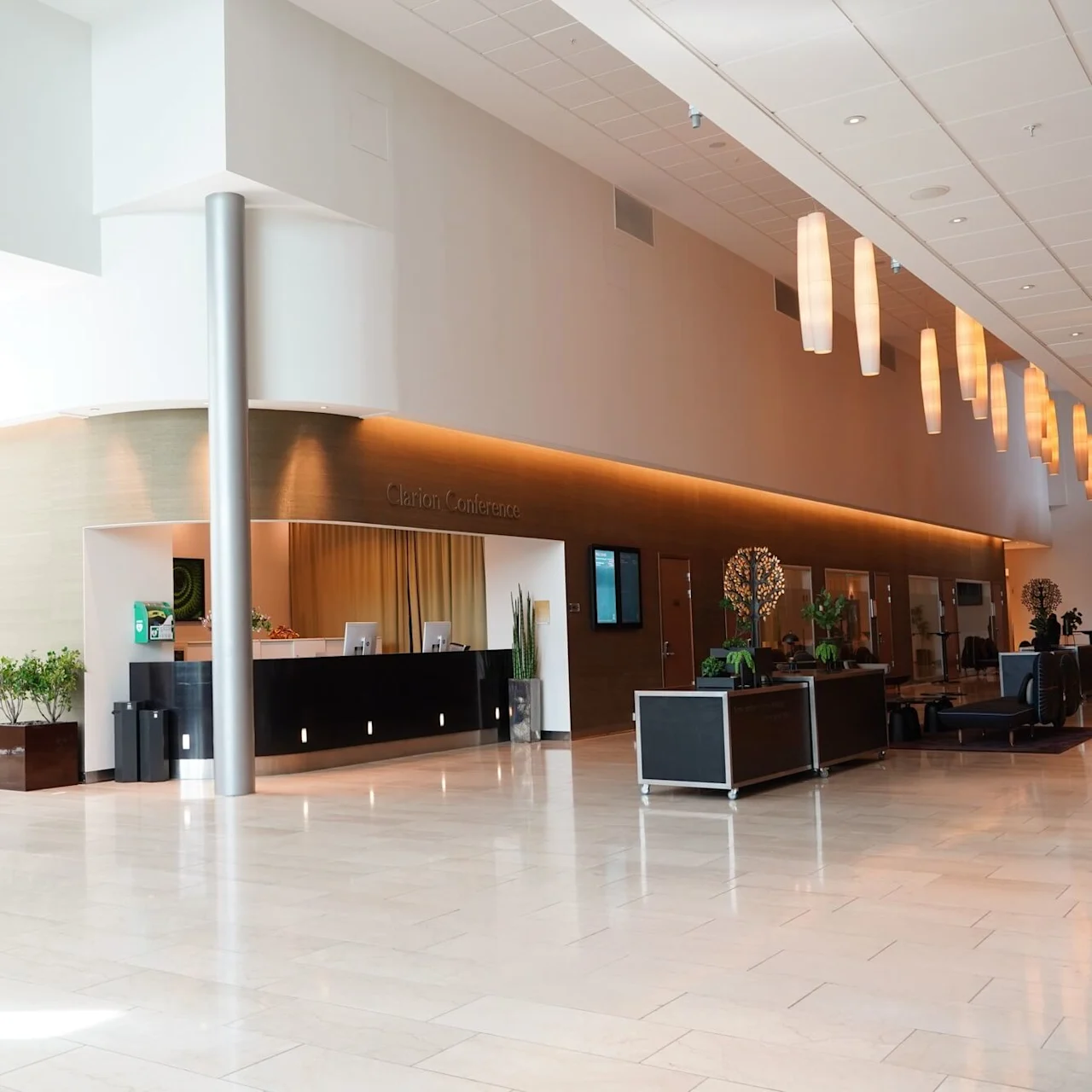 Reception desk stands unmanned, surrounded by potted plants and modern decor in an expansive, brightly lit hotel lobby with hanging lights. Text: "Clarion Conference."