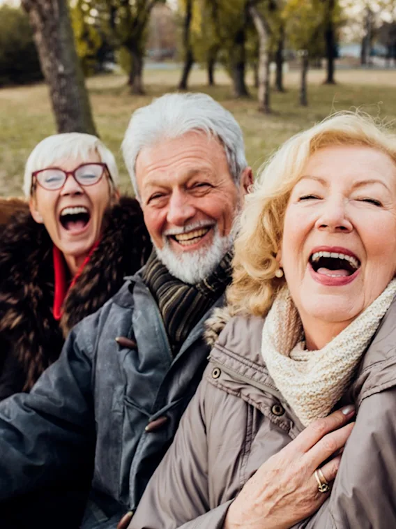 Neljä iloista vanhempaa aikuista nauraa ja hymyilee ottaessaan selfietä ulkona puistossa.